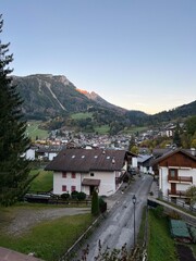 Fototapeta premium A scenic view of Moena, Italy, with mountains in the background, houses, and a street leading uphill during sunrise, showcasing an autumn landscape.