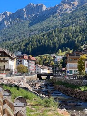 A picturesque view of the town of Moena nestled in the mountains, with houses lining a river, surrounded by lush greenery and towering trees.