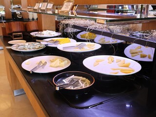 A hotel buffet station features white plates of sliced cheese, wedges, and tongs, with a mirrored display and decorative twine. Ideal for food, buffet, catering, and hospitality imagery.