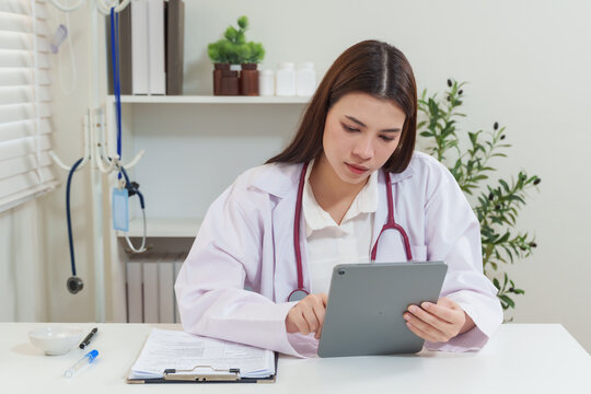 Asian female doctor holding tablet and checking patient details during telemedicine consultation, healthcare professional offering remote medical advice through online technology