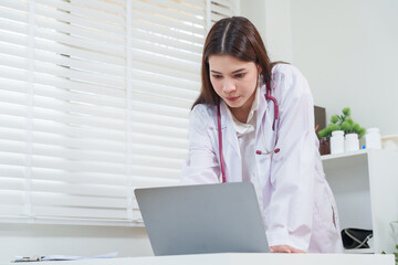 Asian female pediatrician working in clinic leaning over laptop checking medical data patient records representing professionalism dedication healthcare expertise and treatment consultation care