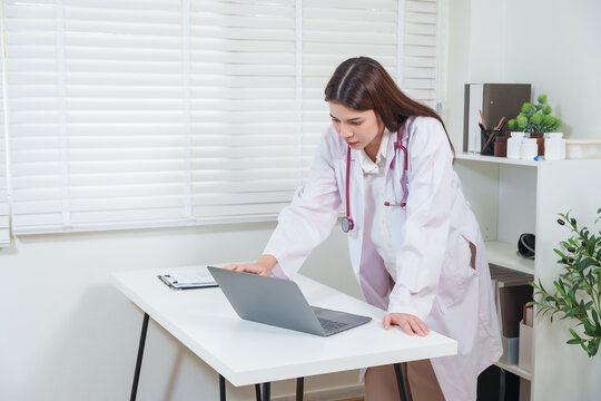 Asian pediatrician standing beside desk in clinic focused on laptop analyzing patient information showing healthcare professionalism dedication treatment consultation and medical expertise concept