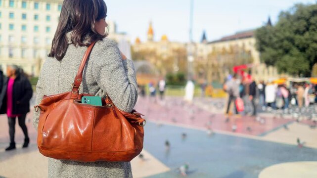 Woman walking through a busy barcelona city square, her handbag open with a mobile phone visible, symbolizing the risk of theft and the importance of traveler caution in an urban environment