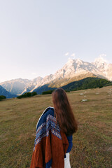 Fototapeta premium Woman back view standing in meadow, looking at snow-capped mountains and wide landscape. Nature travel scene with long hair, coat draped over shoulder and hiking mood.