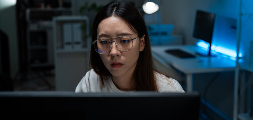 Serious Asian woman with glasses working on computer in dark office. Representing programmer, IT specialist, and modern professional digital work. Software developer programming coding for application
