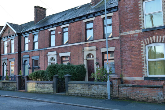 A row of red brick terraced houses are shown from across the street. Each house has an arched entrance and a dark roof in Heywood - Greater Manchester - UK