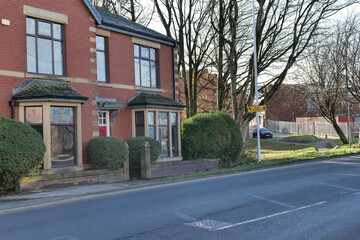 A red brick house with two bay windows sits along a street. Green hedges accent the front of the building with bare trees in the background in Heywood - Greater Manchester - UK