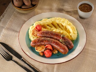 Sizzling pork sausages rest beside fluffy mashed potatoes and caramelized tomatoes on a vibrant plate, evoking the warmth of home-cooked comfort food during chilly days