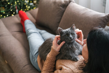 Woman in cozy sweater is relaxing on sofa petting fluffy gray cat on her chest with decorated Christmas tree in blurred background. Christmas comfort, pet love, festive season, relaxing winter weekend © Elen Nika