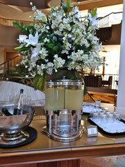 Luxurious hotel lobby event setup featuring a polished beverage station, a tall white flower bouquet, a lemonade dispenser, and a champagne ice bucket for guests.