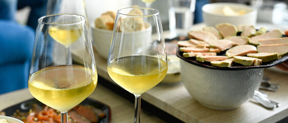 Scène d'apéritif composé de plusieurs verres de vin blanc et d'assiettes de foie gras et saumon fumé, sur des tables basses dans un salon de jour, format bannière