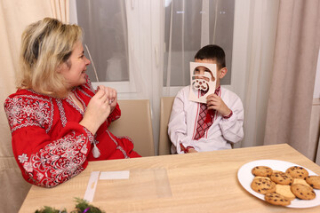 A woman and a boy in embroidered shirts play with paper masks at the table, creating a festive mood before Christmas and New Year.