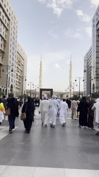 Pilgrims (Hujjaj/Mutamireen) walking in white and black attire down a city street lined with tall buildings towards the iconic minarets of the Prophet's Mosque (Masjid Nabawi) in Medina.