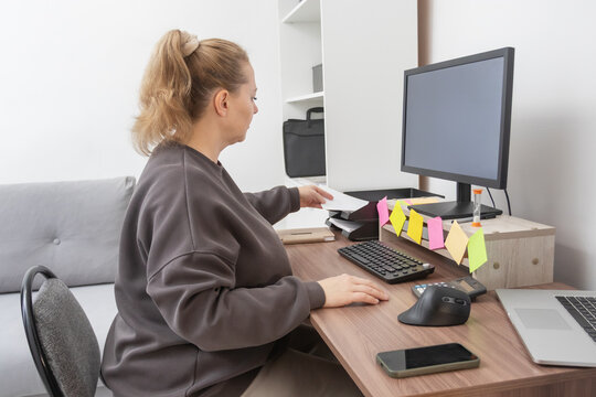 Caucasian woman working from home, planning and writing notes at a wooden desk with a computer monitor, showcasing a casual remote office setup.