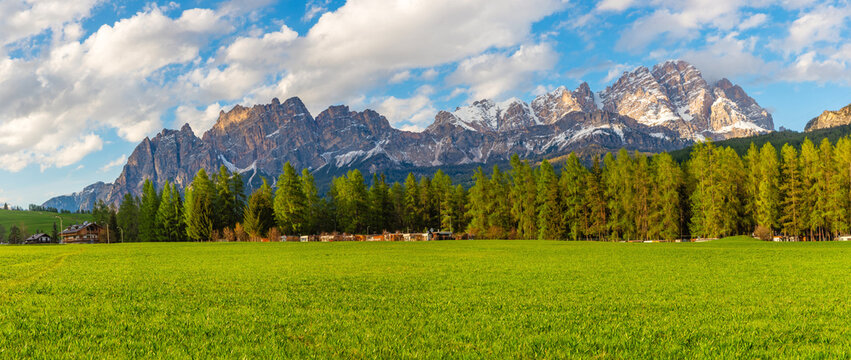 Wide panoramic view of Cortina d'Ampezzo with lush green field, pine forest and snow covered Dolomiti Alps mountains. Italian Dolomites landscape in Belluno region, Italy. Travel destination