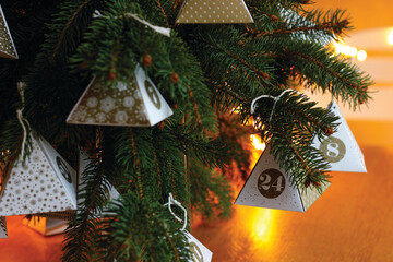 Close-up of a small paper pyramid with the number 24 hanging from a spruce branch, Christmas lights in the background. Sustainable advent calendar. Concept for Christmas Eve