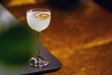 An elegant, pale cocktail in a stemmed coupe glass, garnished with a dried citrus wheel, on a bar counter with shallow depth of field