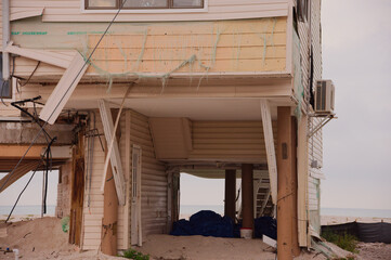 A partially elevated Beach House Damaged by Storm With Exposed Framework and Sand Underneath on the Shoreline in Treasure Island, FL. Sagging deck, and sand-filled ground. The scene captures coastal e