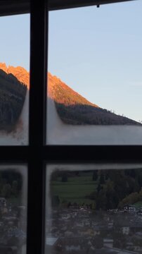 A stunning view of the mountains surrounding Moena, Italy, captured through a window at sunrise, with the peaks bathed in golden light and the landscape below.