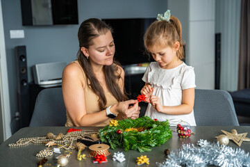 Fototapeta premium Mother and her little daughter decorating a Christmas wreath together at home. Strengthening family bonds through shared festive traditions and creative seasonal activities.