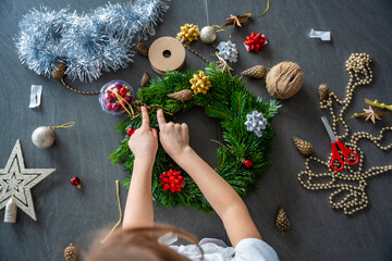 Fototapeta premium Little girl decorating a Christmas wreath while sitting at the table, top view. Capturing a hands-on festive activity from an overhead perspective at home.