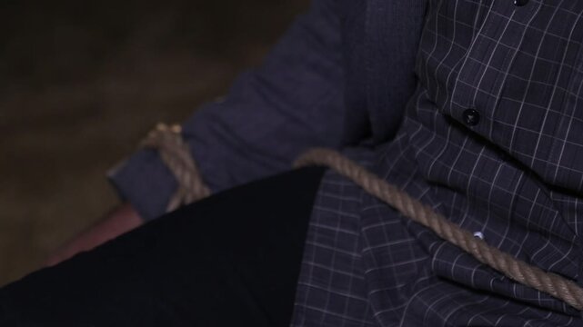 Close up of hostage's hands bound with tape to a chair while struggling to escape. Followed by hands tied with rope around a wooden post in a dark room.

