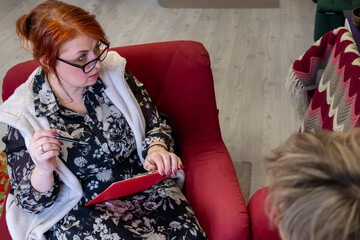 Woman with red hair wearing glasses is seated on a red couch, holding a tablet and pen, engaged in conversation, showcasing a collaborative and creative environment