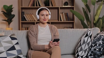 Caucasian woman sitting on sofa listening to music in white headphones. Holding smartphone and nodding head to rhythm. Calm female enjoying favorite playlist at home surrounded by books and plants.