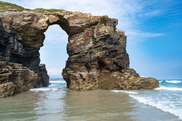 Rock arch formation at As Catedrais Beach in Ribadeo Galicia, Spain, with coastal cliffs, gentle waves, and a bright blue sky creating a stunning seascape