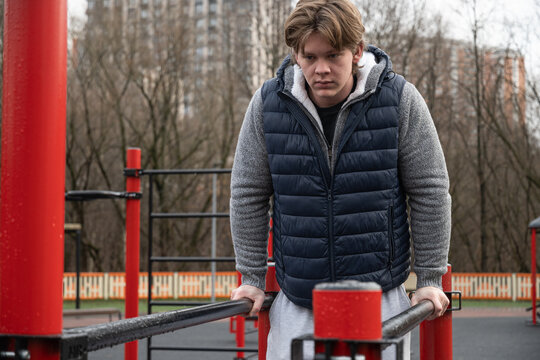 Focused man exercise on parallel bars in an urban outdoor gym after rain. Dressed warmly, concentrating on fitness routine amidst city backdrop, emphasizing strength and determination. Daily routine