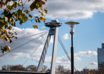 Street lamp in the foreground with the SNP Bridge and its UFO observation tower in Bratislava, Slovakia, captured on a sunny day with clouds and autumn leaves.