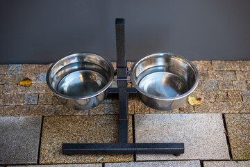 Two stainless steel bowls on a metal stand placed outdoors, used for providing food and water for animals.