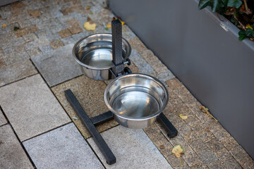 Two stainless steel bowls on a metal stand placed outdoors, used for providing food and water for animals.