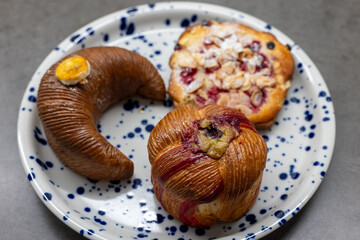 Plate with artisanal pastries including a banana-shaped banana cake, a fruity muesli cheesecake pastry, and a pistachio-filled pastry, perfect for breakfast or coffee break.