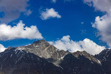 Obraz premium Rugged, snow-dusted mountain peaks rise against a vibrant blue sky, dotted with fluffy white clouds under bright, natural daylight.