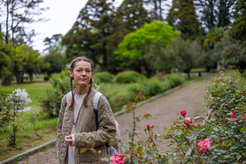 A woman with braided hair stands on a garden path, wearing a quilted jacket and carrying a backpack. Pink roses bloom in the foreground, with lush green trees and soft, diffused lighting in the backgr