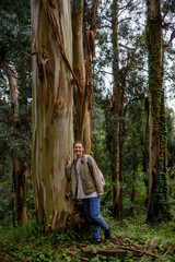 A smiling woman leans against a large eucalyptus tree with peeling bark in a lush, green forest. Soft, diffused daylight filters through the canopy, highlighting the natural textures and earthy tones