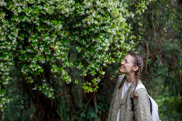 A smiling woman with a braided hair stands in a lush green forest, gazing up at a flowering bush with white blossoms. Soft natural light illuminates the scene.