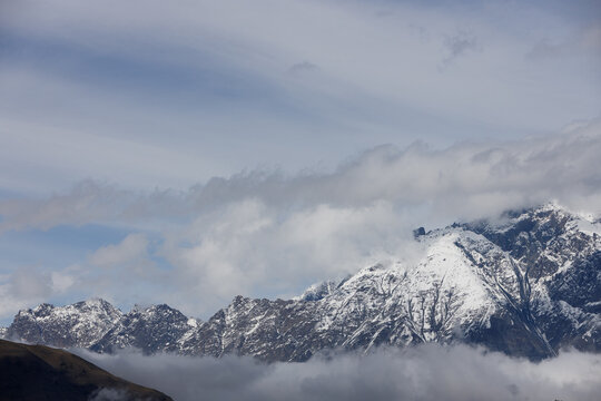 Snow-capped mountains rise above swirling clouds under a soft, overcast sky, with muted blues and grays dominating the dramatic, misty landscape.