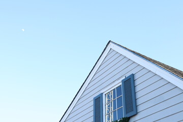 The upper gable section of a light blue-sided house, featuring a window with dark blue shutters, is framed against a pale blue daytime sky with a visible sliver of the moon.