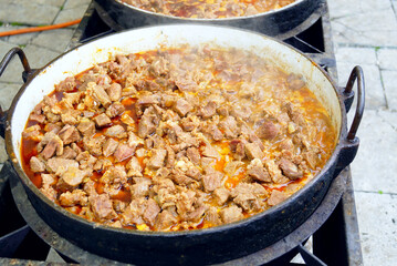 Large cast iron pan filled with tender meat pieces in thick tomato-based sauce on outdoor gas stove. Illustrates outdoor cooking experience, traditional goulash recipe, and rustic meal preparation.