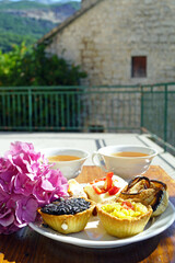 Elegant tea service with assorted tartlets and hydrangea blooms on an outdoor terrace with mountain view. Illustrates alfresco dining experience, Mediterranean lifestyle aesthetic, artisanal pastry.