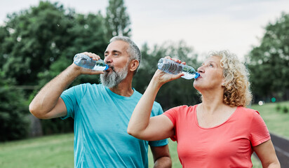 Naklejka premium Smiling active mature mid aged couple jogging exercising and having fun and laughing together taking a break drinking water in the park