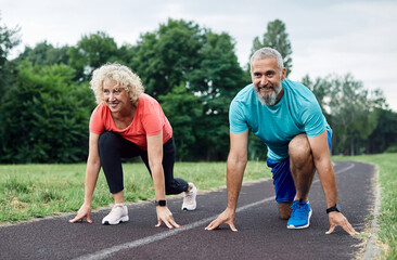 Smiling active mid aged couple standing in start position,  jogging running and walking doing fitness in the park