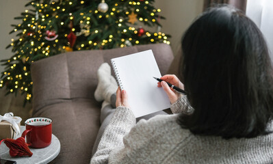 Woman in cozy sweater writing in blank spiral notebook while relaxing on sofa with festive Christmas tree in background. Planning, resolutions, to-do list, wish list, mockup, shopping list