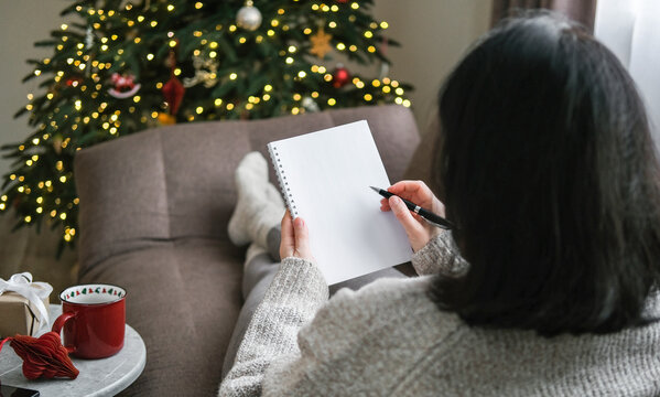 Woman in cozy sweater writing in blank spiral notebook while relaxing on sofa with festive Christmas tree in background. Planning, resolutions, to-do list, wish list, mockup, shopping list