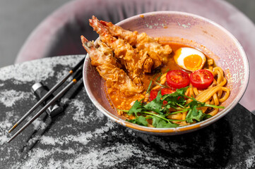 A bowl of spicy tempura shrimp ramen with noodles, a soft-boiled egg, cherry tomatoes, and cilantro, on a marble table with chopsticks