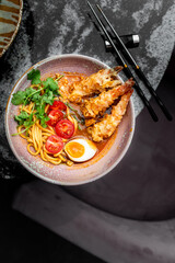 A close-up, high-angle photo of vibrant tempura shrimp ramen in a textured bowl with noodles, a soft-boiled egg, cherry tomatoes, and cilantro on a dark stone table