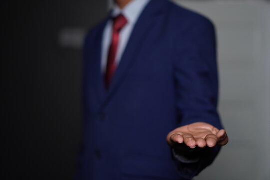 Businessman in a blue suit and red tie extends his open hand forward in a gesture of offering or presentation in a dark setting