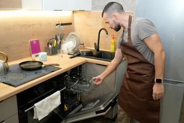 A man in an apron is busy in the kitchen preparing dinner. He is loading a dishwasher with clean dishes next to a stove and various cooking utensils on the counter